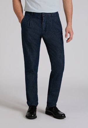 Uomo che indossa pantaloni sartoriali blu scuro con pieghe, mocassini in pelle nera e una camicia bianca infilata, in piedi contro uno sfondo semplice.