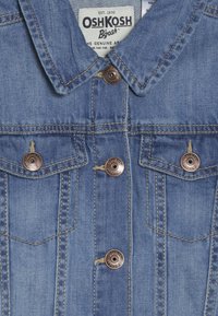 Džínová bunda s bledě modrou úpravou, špičatým límcem a měděně tónovanými knoflíky s vyrytým vzorem. Dvě náprsní kapsy s knoflíkovými uzávěry.