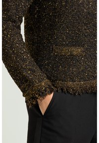 Sort tekstureret sweater med guldtrådsaccenter. Har frynser på kanterne og en afslappet pasform. Bruges over sorte bukser, der viser minimal detaljer.