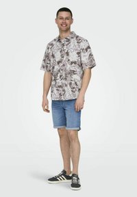 Kurzärmliges Blumenshirt mit einem beigen und grauen Muster, kombiniert mit hochgekrempelten Jeansshorts und dunklen Turnschuhen mit weißen Streifen.