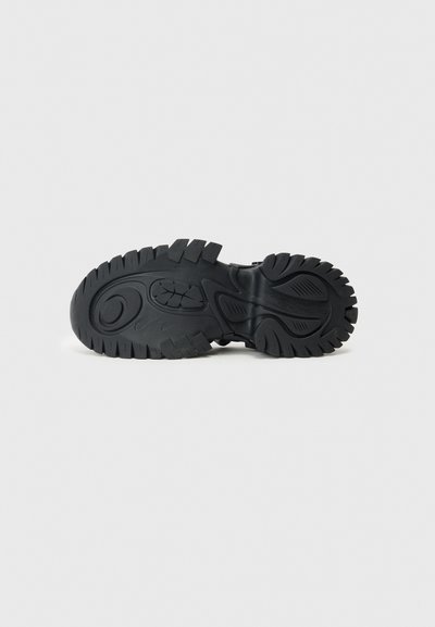 Black rubber shoe sole with thick, uneven tread pattern and embossed logo. Texture is smooth with a durable design for traction.