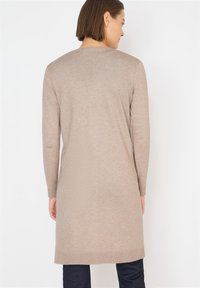 Dlouhý béžový cardigan vyrobený z měkkého pleteného materiálu. Má dlouhé rukávy a kulatý výstřih, s rovnými lemy sahajícími do poloviny stehna.