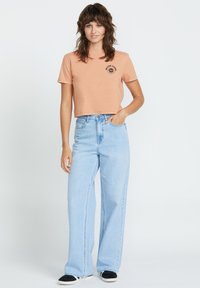 Jasno brzoskwiniowy, krótki t-shirt z czarną grafiką słońca, zestawiony z jasnoniebieskimi, szerokimi spodniami z wysokim stanem i czarnymi butami wsuwanymi.