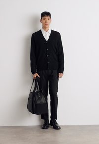 Cardigan negro con botones, camisa blanca con cuello, pantalones negros y zapatos. Sostenido un bolso gris con acentos negros. Diseño simple y moderno.