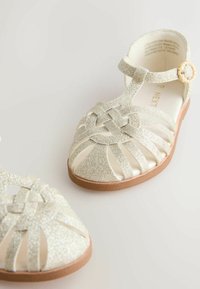 Glittery silver sandals with a woven design, open toes, ankle strap, and decorative buckle. Light sole with a textured finish.