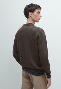 Giovane uomo con capelli ricci corti che indossa un maglione marrone sopra una camicia scura e pantaloni beige, rivolto di spalle con la testa inclinata di lato.