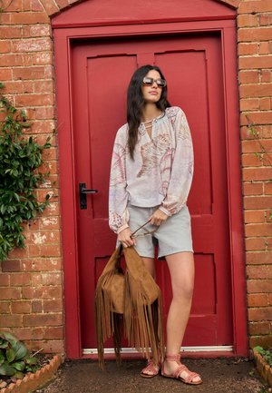Woman wearing floral long-sleeve blouse, gray shorts, and sunglasses stands in front of red door holding a brown fringed handbag.