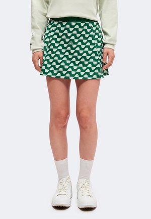 Personne portant une jupe à motif ondulé vert et blanc, une chemise blanche à manches longues, des chaussettes blanches et des chaussures blanches, debout, les bras détendus.