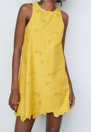 Vestido amarillo sin mangas con patrón de encaje floral y dobladillo festoneado, llevado por una persona con piel oscura y pendientes con cuentas.