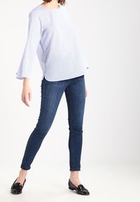 Camicia a righe blu e bianca a maniche lunghe con un taglio morbido, abbinata a jeans slim blu scuro e scarpe nere, in piedi su uno sfondo neutro.