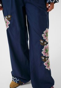 Pantaloni largi de marină cu broderie florală în nuanțe de roz, verde și maron. Materialul pare ușor; tivul are un accent cu un model.