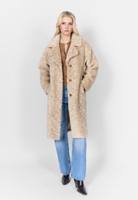 Beige nepbontjas met een notar collar, knopen aan de voorkant en zijzakken, gecombineerd met blauwe flare jeans en donkere puntige laarzen.