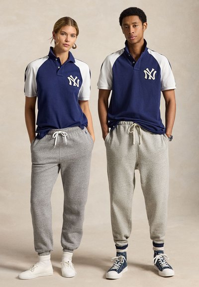 Mees ja naine seisavad kõrvuti, kandes tumesiniseid ja valgeid New York Yankees polo-särke ning halli dressipükse, käed taskutes.