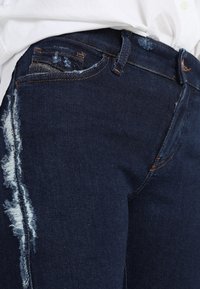 Jeans de mezclilla azul oscuro con un detalle desgastado en una pierna, que presentan un diseño clásico de cinco bolsillos y costuras en contraste.