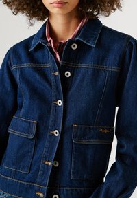 Σκούρα μπλε τζιν τζάκετ με αντίθετες πορτοκαλί ραφές, μπροστινές τσέπες και ασημένια κουμπιά. Διαθέτει γιακά και άνετη γραμμή.