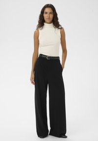 Vrouw met lang krullend haar, gekleed in een mouwloze crème coltrui, zwarte wijde broek, zwarte riem en zwarte schoenen, staand tegen een witte achtergrond.