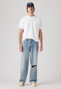 Νεαρός άνδρας που φορά μπλε σκούφο μπέιζμπολ, απλό λευκό μπλουζάκι, ανοιχτό μπλε τζιν με σκισίματα και μπεζ αθλητικά παπούτσια, στέκεται μπροστά σε απλό φόντο.