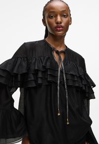 Bluză neagră cu multiple straturi de volane, mâneci lungi și un detaliu cu fundă la gât, accentuat cu elemente metalice și mărgele mici.