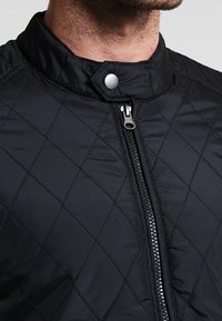 Giacca nera trapuntata con colletto alto, bottone a pressione argento e zip frontale. Tessuto liscio con cuciture a motivo diamantato.
