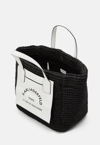 Svart flätad tote-väska med vita läderhandtag och en rektangulär varumärkeslapp med text. Rymligt inre med detalj av dragkedjeficka.