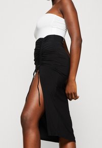 Svart högmidjad rynkad kjol med slits vid låret, bärs med vit bärbar topp, sidvy av stående person mot en enfärgad bakgrund.