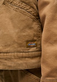 Brun kanvasjakke med synlig Dickies-logo-badge, teksturert stoff og rett snitt laget med lommedetaljer.