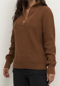 Femme portant un pull marron en maille avec une demi-fermeture éclair dorée et un pantalon noir, debout devant un fond uni.