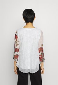 Blusa blanca con tejido transparente, mangas tres cuartos con bordados florales rojos y un ligero curvado en el dobladillo. Ligera y texturada.