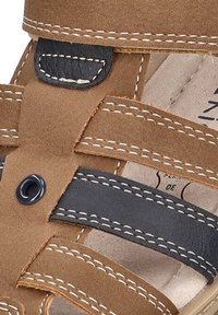 Sandaña de ante marrón con correas de cuero negro, costuras blancas y un detalle de ojal circular. Diseñada para la comodidad con una plantilla texturizada.