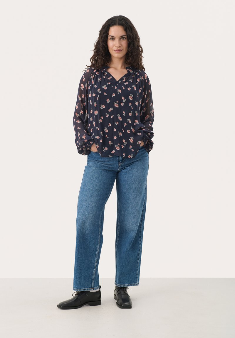 Navy-Bluse mit kleinem Blumenmuster, langen Ärmeln und V-Ausschnitt. Kombiniert mit weit geschnittenen blauen Jeans und schwarzen Schuhen. Glatte Texturen sichtbar.