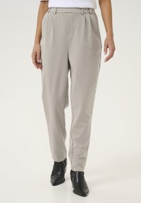 Pantalons taillés gris clair à taille haute, avec des plis sur le devant et une texture lisse, associés à des bottines noires.