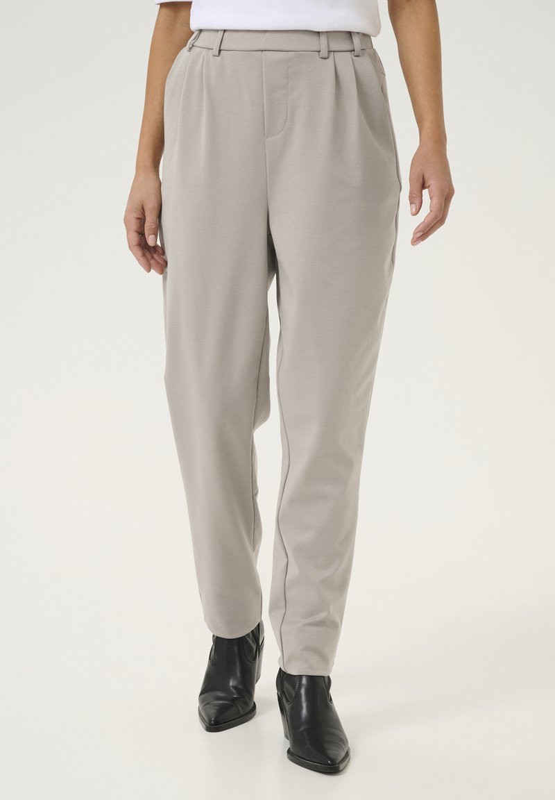 Pantalons taillés gris clair à taille haute, avec des plis sur le devant et une texture lisse, associés à des bottines noires.