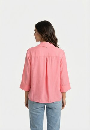 Femme aux longs cheveux foncés portant un chemisier rose ample à manches trois-quarts et un jean bleu clair, dos à l'appareil photo.