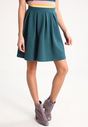 Kvinde iført teal plissert nederdel, flerfarvet stribet top og grå ruskindsankelstøvler stående på hvid baggrund.