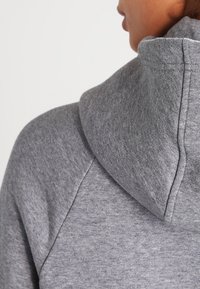 Grå sweatshirt med mjukt, strukturerat tyg, utrustad med stor huva och raglanärmar, som visar sömmar i detalj och en avslappnad design.