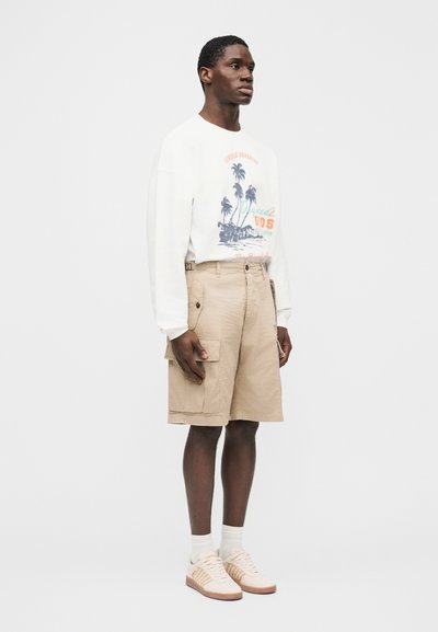 Κρεμ φούτερ με γραφικό σχέδιο φοίνικα και κείμενο, συνδυασμένο με μπεζ cargo σορτς με πλαϊνές τσέπες. Μπεζ αθλητικά παπούτσια με πορτοκαλί λεπτομέρειες.
