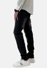 Pantalones vaqueros de mezclilla negra exhibidos en vista lateral, con un corte slim, costuras mínimas y una altura de cintura media, combinados con zapatillas blancas.
