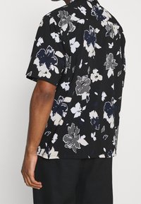 Camisa negra de manga corta con un patrón floral en gris, blanco y azul marino. Corte holgado con un cuello tipo polo y una textura sutil.