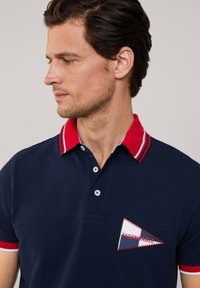 Granatowa koszulka polo z czerwonym paskiem przy kołnierzyku, z trójkątną naszywką z logo i trzema guzikami, wykonana z bawełny o teksturowanym wykończeniu.