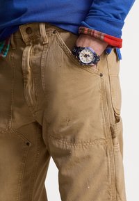 Beige arbetsbyxor med fickor, lätt slitage, bärs med en blå långärmad skjorta. En svart klocka har en vit urtavla och blå accenter.