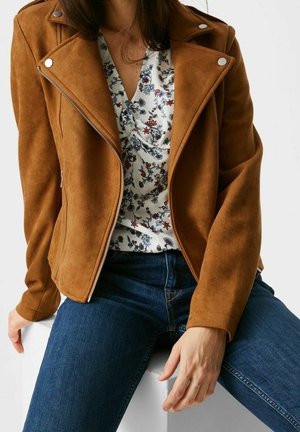 Kvinde iført en brun ruskindjakke over en hvid blomstret bluse og blå jeans, siddende med den ene hånd hvilende på låret.