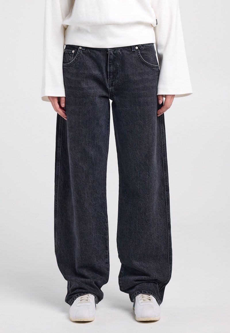 Vaqueros de mezclilla negros de pierna ancha con una textura desgastada, con herrajes plateados, bolsillos laterales y un ajuste relajado, combinados con zapatillas blancas.