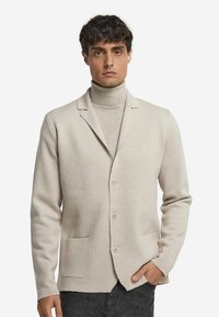 Mann, der eine beigen, geknöpften Cardigan über einem passenden Rollkragenpullover und dunklen Hosen trägt, steht mit einer Hand in der Tasche vor einem weißen Hintergrund.