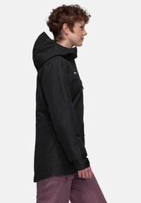 Schwarze wasserdichte Jacke mit Kapuze, Frontreißverschluss und Seitentaschen. Zeigt eine entspannte Passform und strukturierte Stoff. Kombiniert mit rosa Hosen.