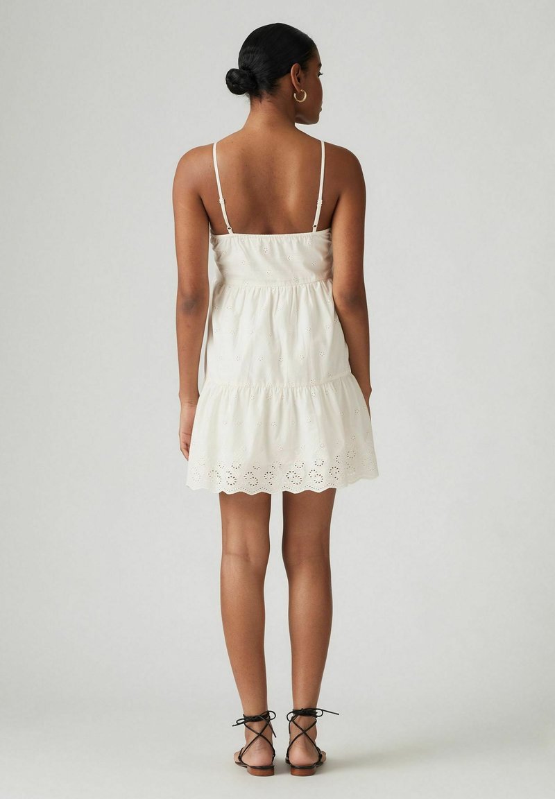 Vestido de verano blanco de algodón con tirantes ajustables, diseño en capas y bordado de ojales en el dobladillo. Combinado con sandalias negras con tiras.