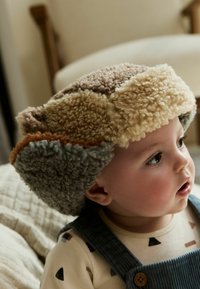 Multicolored fleece hat with a patchwork design, featuring shades of beige, brown, and gray. Soft texture with ear flaps.
