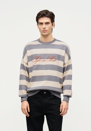 Muž nosiaci béžový a sivý horizontálne pruhovaný sveter s oranžovým vyšívaným nápisom "Lyle and Scott", kombinovaný s čiernymi nohavicami.