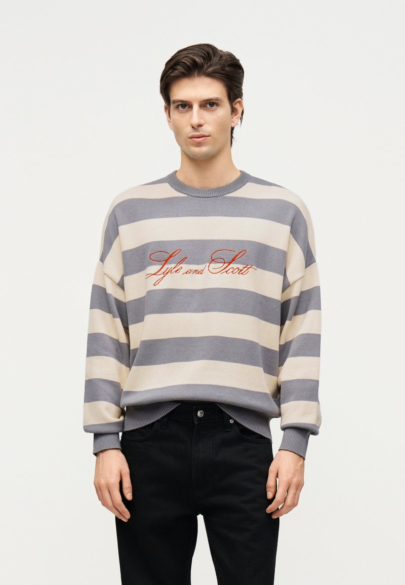 Άνδρας που φοράει μπεζ και γκρι οριζόντια ριγέ πουλόβερ με κεντημένο το "Lyle and Scott" σε πορτοκαλί, συνδυασμένο με μαύρο παντελόνι.