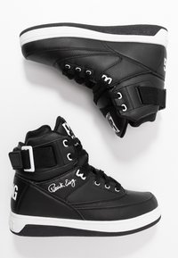Baskets montantes noires avec semelles intermédiaires blanches, lacets, sangles à la cheville et la signature "Patrick Ewing" sur le côté.