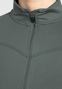 Chaqueta gris con cremallera y cuello alto, fabricada en un tejido suave y texturizado, con costuras contrastantes a lo largo de las costuras.